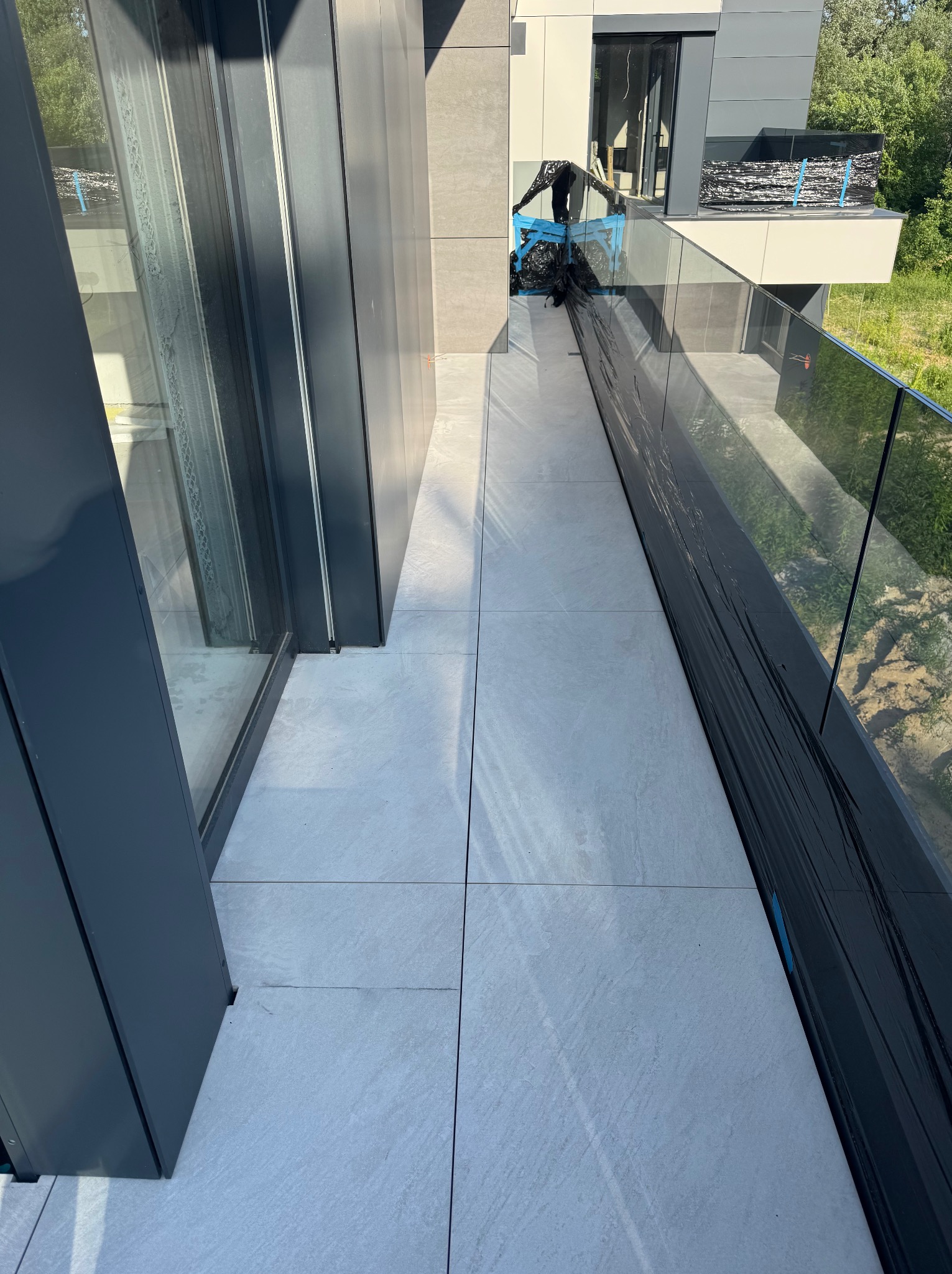 Nowoczesny balkon z szarą, kwadratową płytką w Warszawie. Balustrada szklana, widok na zieleń. Częściowo zabezpieczony folią, przygotowany do prac wykończeniowych.