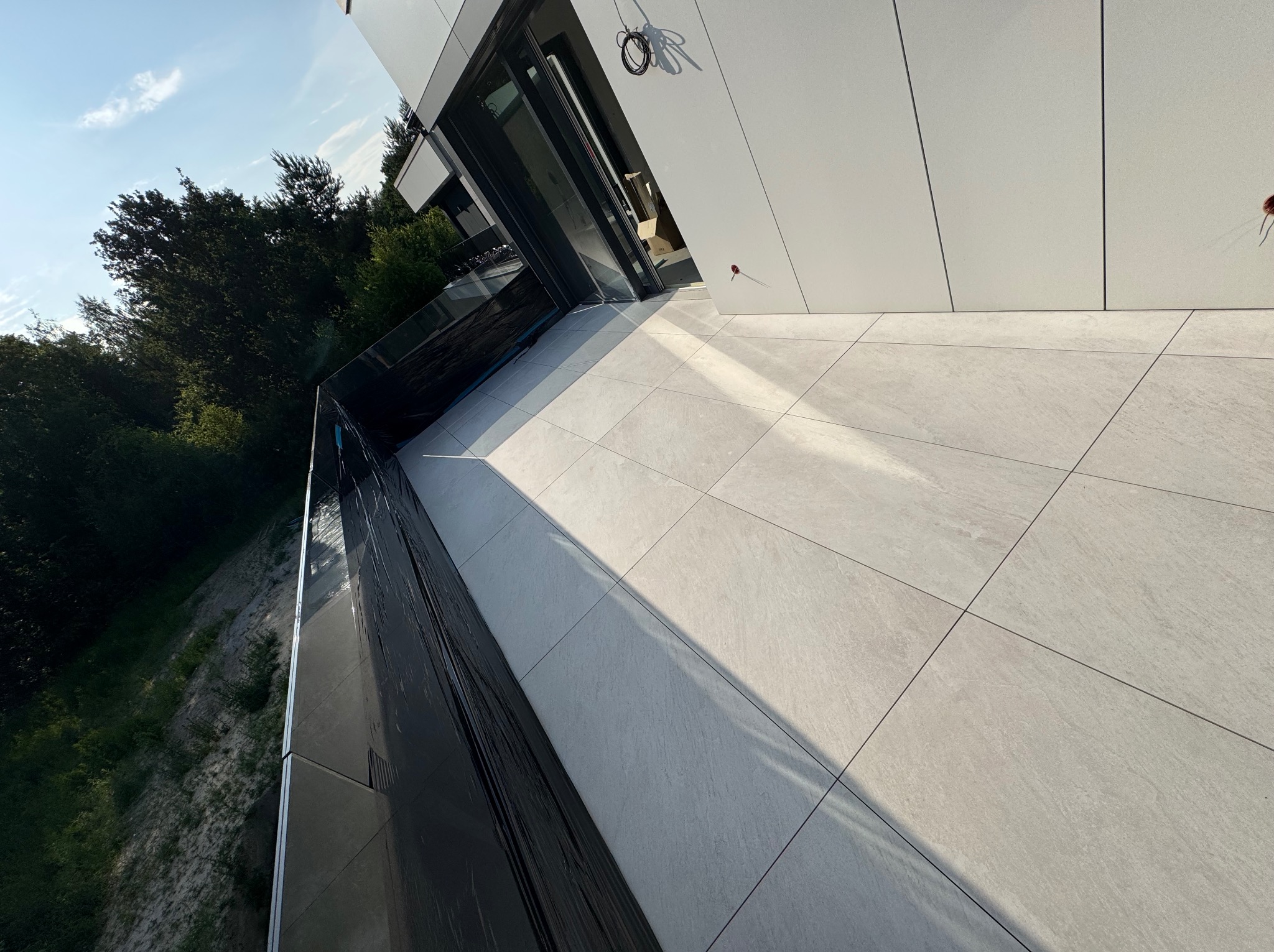 Nowoczesny taras z jasnoszarej, kwadratowej płytki, z widokiem na zieleń. Eleganckie wykończenie balkonu z przeszkloną balustradą i fragmentem elewacji budynku.