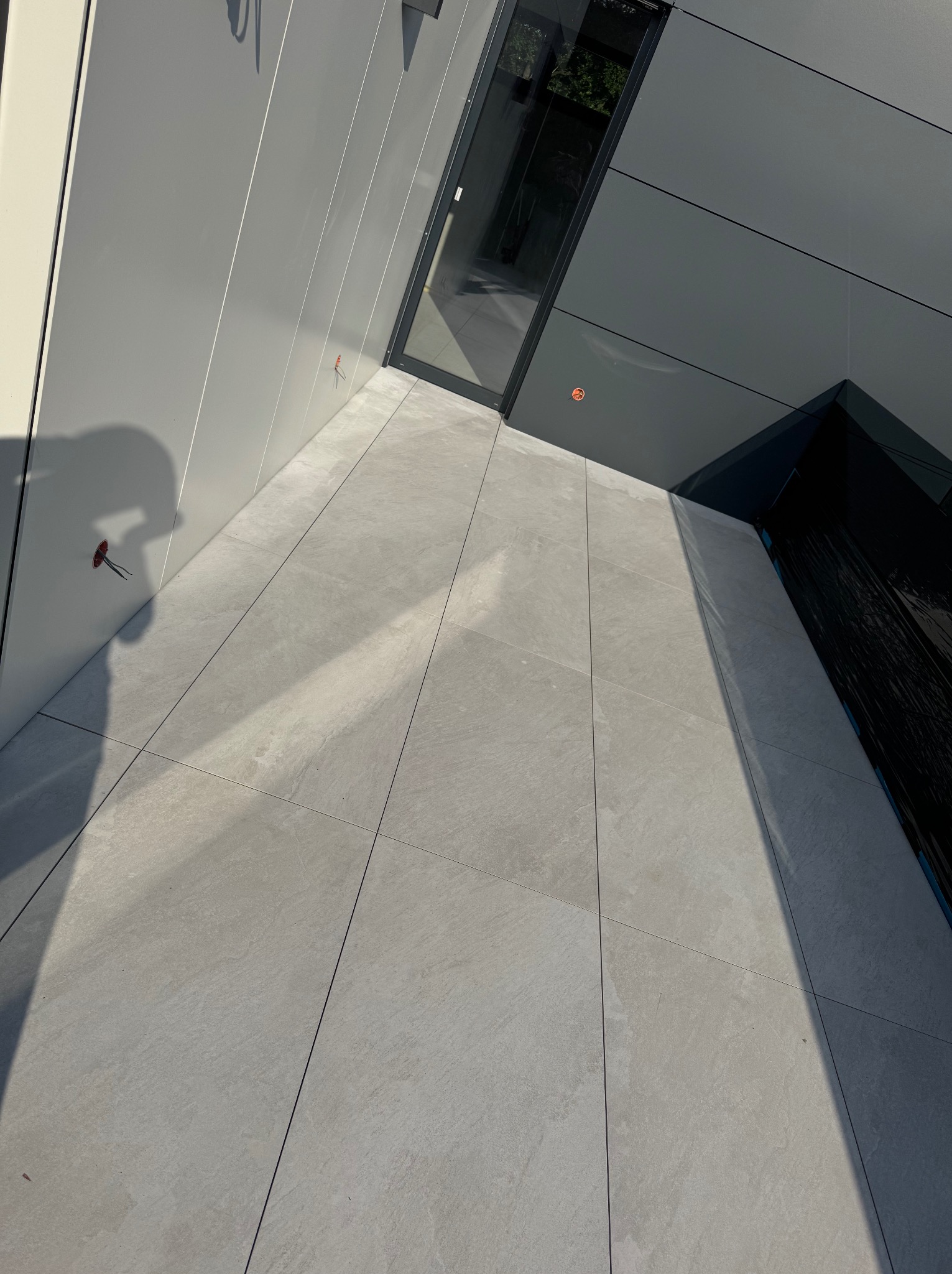 Nowoczesny taras z szarymi płytkami, widok pod kątem. Widoczne drzwi balkonowe i fragment ściany. Cień osoby w lewym rogu. Jasne, naturalne światło.