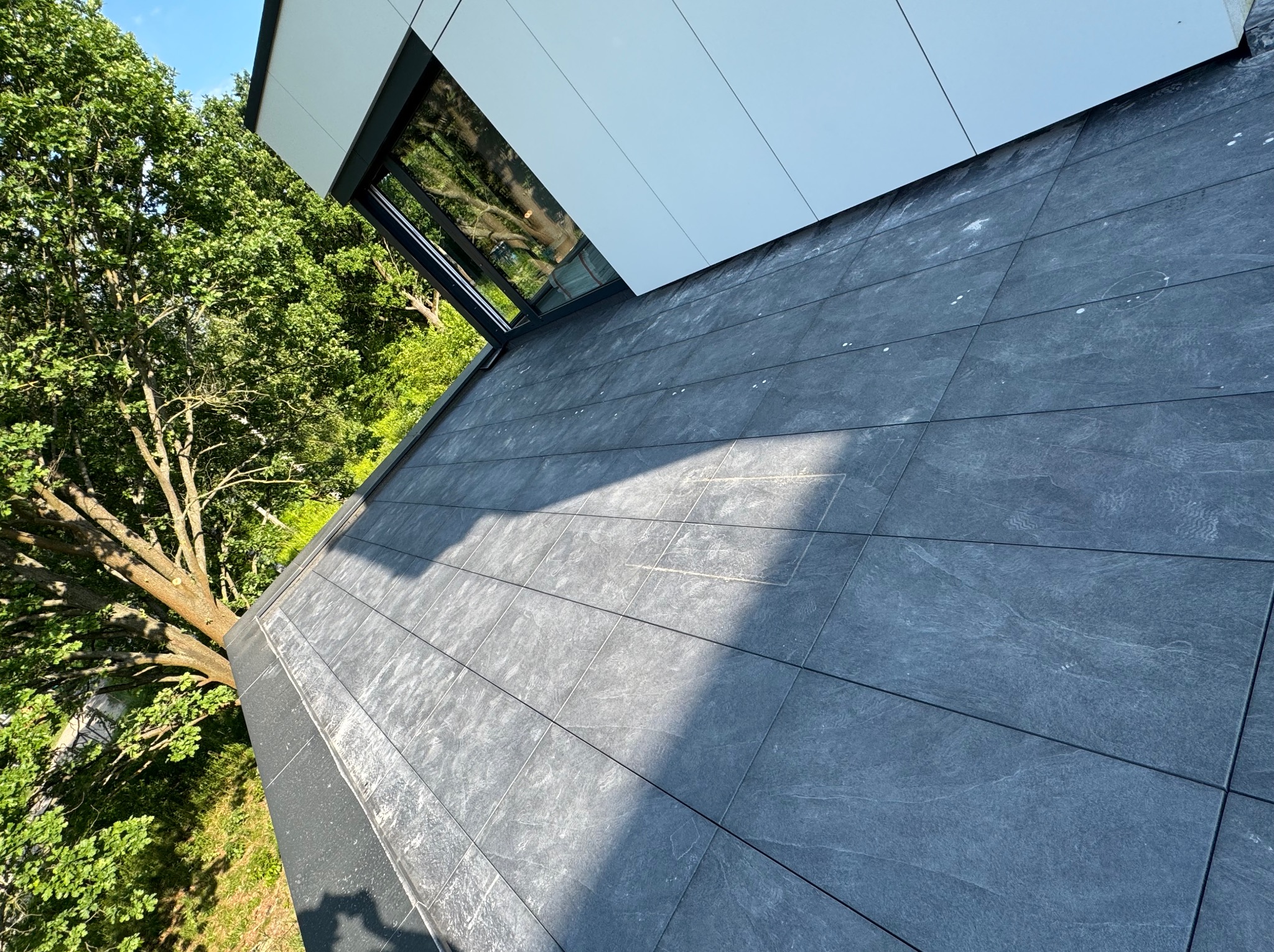 Nowoczesny taras z szarych płyt, przylegający do budynku z oknem. Widok z góry, zieleń drzew w tle. Minimalistyczny design, równa powierzchnia.