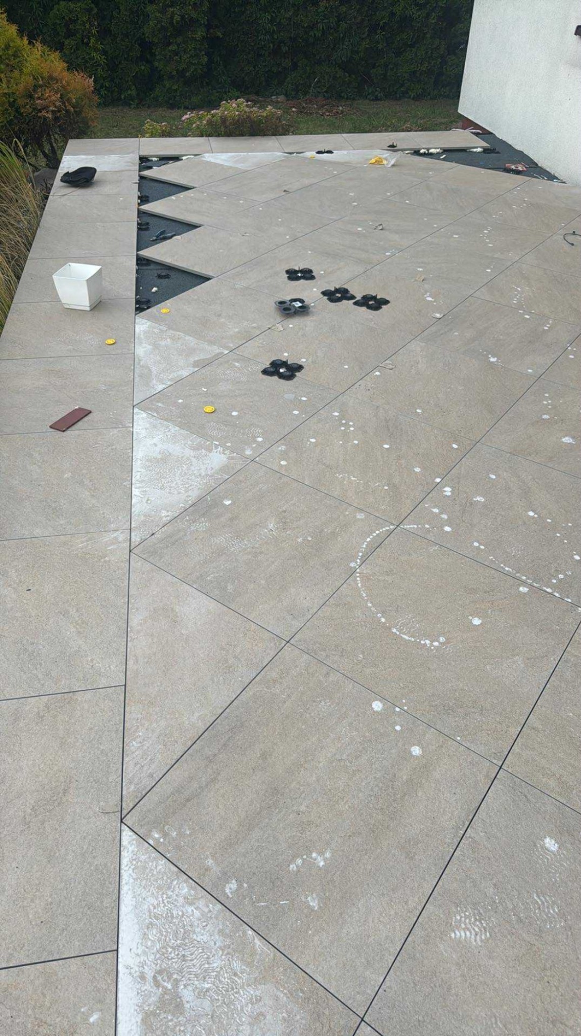 Montaż płytek tarasowych na wspornikach, widoczne elementy mocujące i fragmenty niezakończonej powierzchni. Jasne płytki ułożone w karo, z plamami po zaprawie.