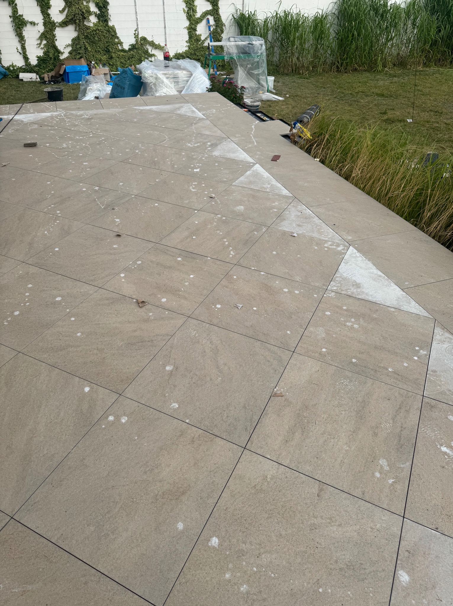 Wykończony taras z jasnych, kwadratowych płyt. Widoczne ślady po fugowaniu. W tle trawa i biały mur porośnięty bluszczem. U góry widoczne materiały budowlane.