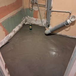 BOB-BUD - Świeżo wylana, mokra wylewka samopoziomująca w łazience podczas remontu, widoczne rury kanalizacyjne i fragmenty ścian z resztkami farby.