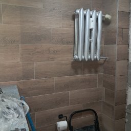 BOB-BUD - Ściana wyłożona płytkami imitującymi drewno, z zamontowanym srebrnym grzejnikiem żeberkowym i akcesoriami łazienkowymi, widoczne narzędzia i drabina.