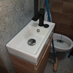 BOB-BUD - Umywalka nablatowa z czarną armaturą i szafką w kolorze drewna, bidet oraz mop w tle na podłodze z wzorzystymi płytkami.