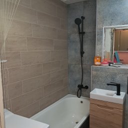 BOB-BUD - Ukończona łazienka z beżowymi płytkami imitującymi drewno na ścianie, czarną armaturą, białą wanną i szafką pod umywalką z drewnianym frontem. Na półce pod lustrem stoją kosmetyki i akcesoria.