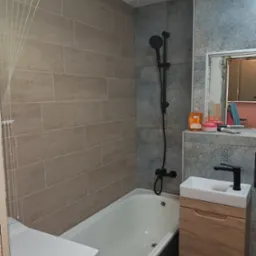 Ukończona łazienka z beżowymi płytkami imitującymi drewno na ścianie, czarną armaturą, białą wanną i szafką pod umywalką z drewnianym frontem. Na półce pod lustrem stoją kosmetyki i akcesoria.