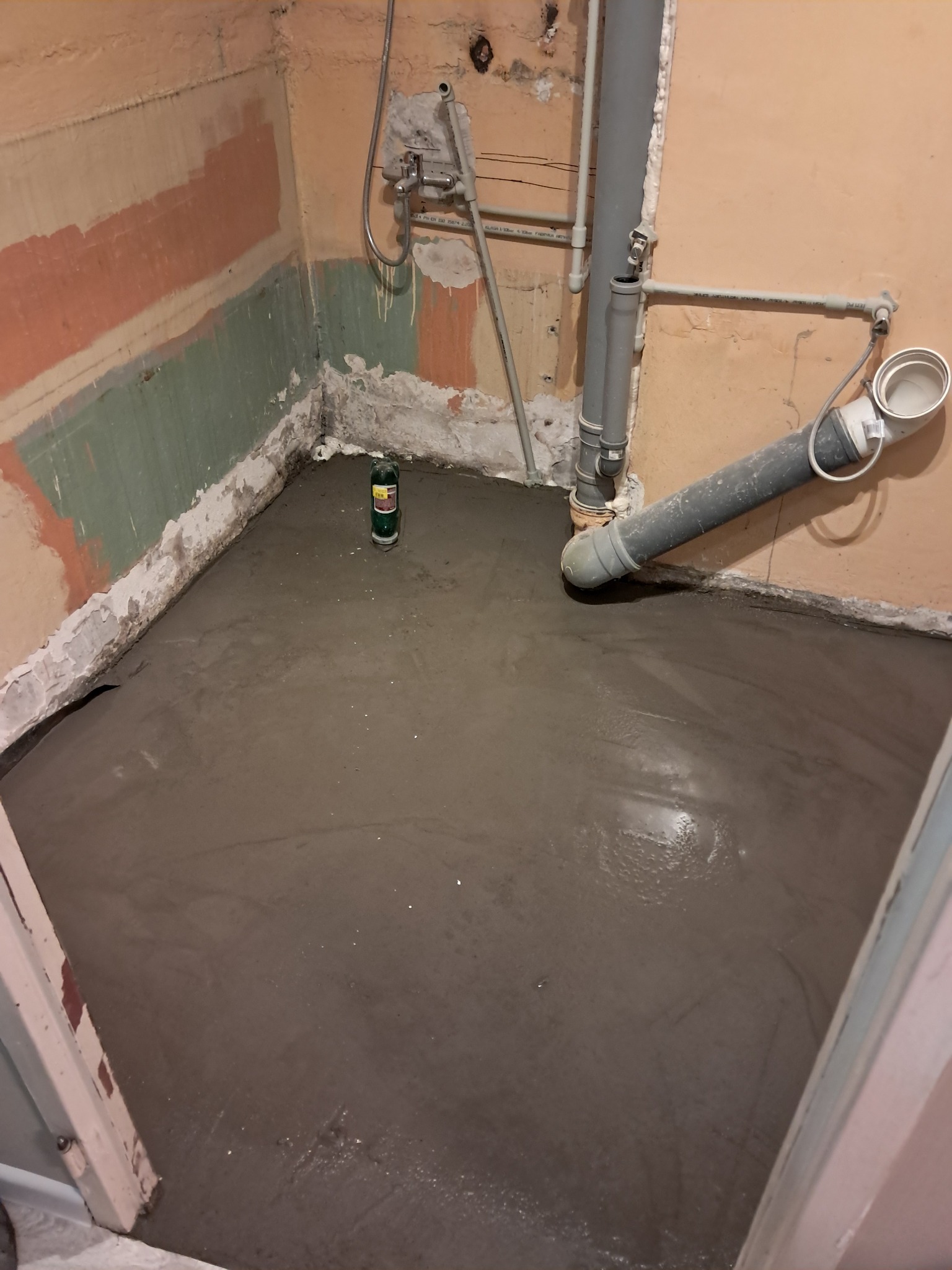 Świeżo wylana, mokra wylewka samopoziomująca w łazience podczas remontu, widoczne rury kanalizacyjne i fragmenty ścian z resztkami farby.