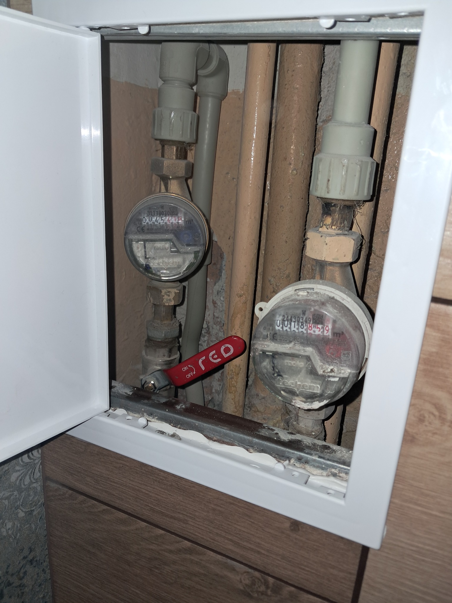 Dwa liczniki wody z czerwonym zaworem odcinającym, widoczne przez otwartą białą szafkę inspekcyjną w łazience z brązowymi rurami.