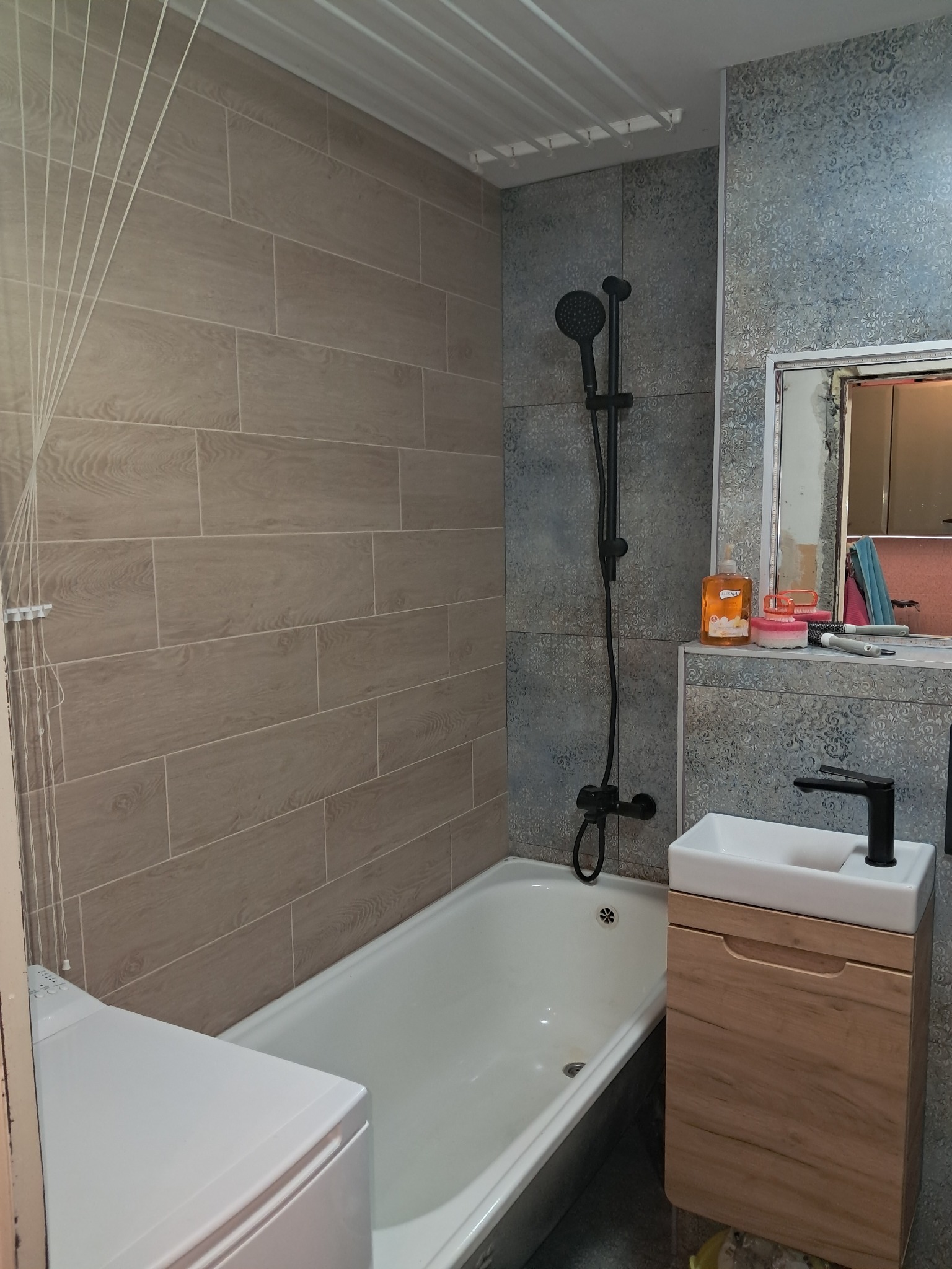 Ukończona łazienka z beżowymi płytkami imitującymi drewno na ścianie, czarną armaturą, białą wanną i szafką pod umywalką z drewnianym frontem. Na półce pod lustrem stoją kosmetyki i akcesoria.