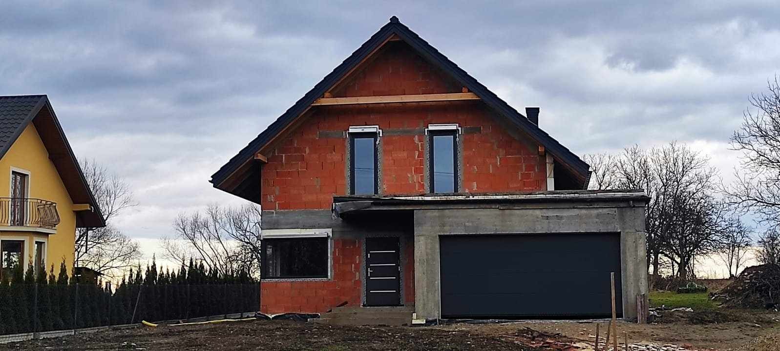 Dom w trakcie budowy z czerwonej cegły, z zamontowanymi oknami i czarną bramą garażową, widoczny w pochmurny dzień.