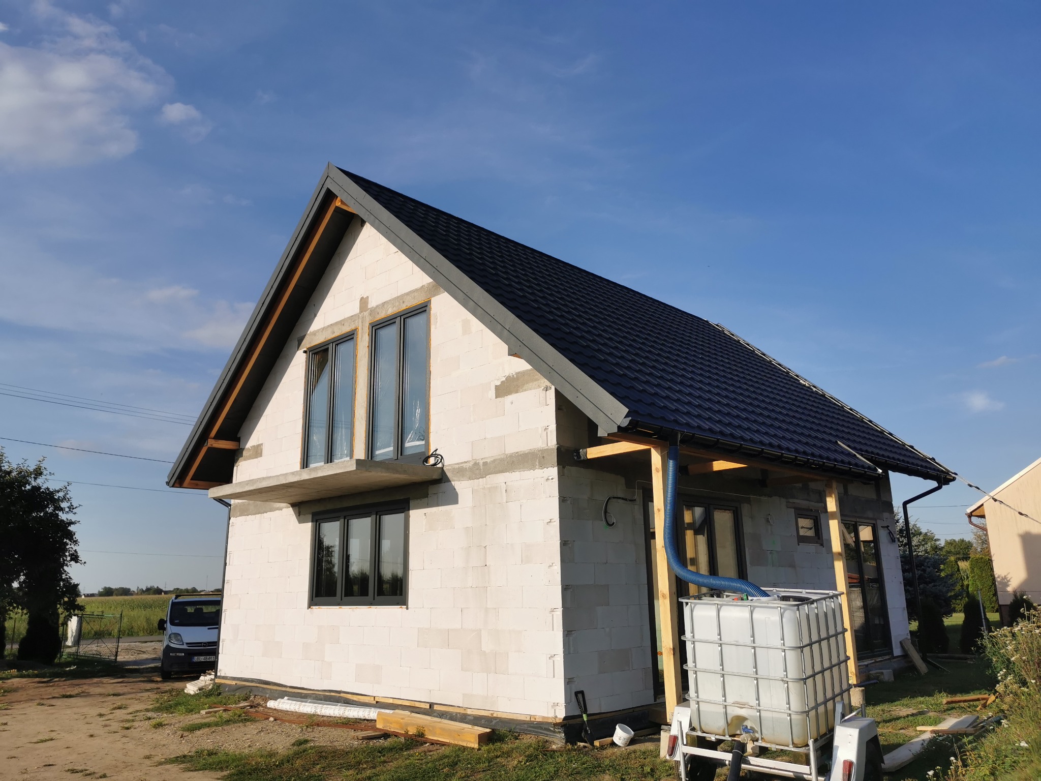 Surowy stan domu jednorodzinnego z białych bloczków, z czarnym dachem i ciemnymi oknami, widoczne elementy konstrukcyjne dachu i zbiornik na wodę z niebieskim wężem na zewnątrz budynku.