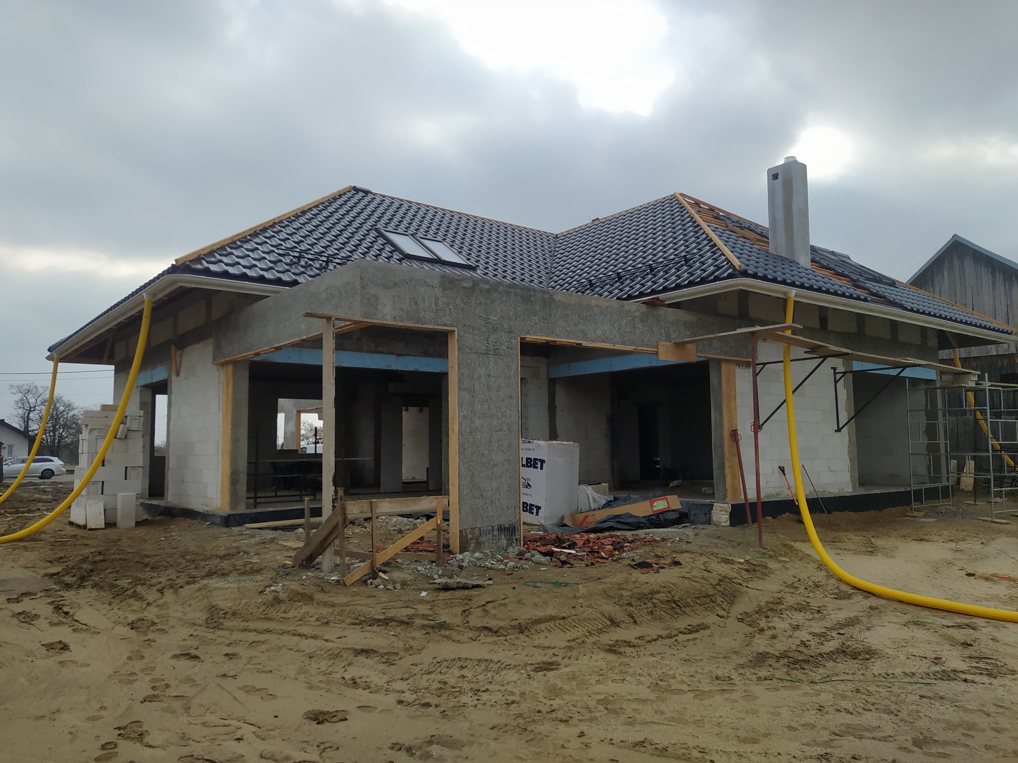 Surowy stan domu jednorodzinnego z czarną dachówką, widoczne otwory okienne i drzwiowe, częściowo otynkowane ściany z betonu komórkowego, żółty wąż na budowie i sterta bloczków Ytong.