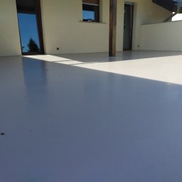 Mikrocement i hydroizolacje - Jasnoszara, błyszcząca powierzchnia tarasu z widocznymi drzwiami balkonowymi, słupem konstrukcyjnym i fragmentem ściany w tle, oświetlona padającym światłem słonecznym.