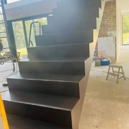 Mikrocement i hydroizolacje - Nowoczesne, ciemnoszare schody wewnętrzne o minimalistycznym designie, z widocznym wykończeniem w stylu industrialnym i surowym betonem na podłodze w tle.
