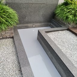 Mikrocement i hydroizolacje - Nowoczesny taras z szarego kamienia i jasną, gładką powierzchnią, otoczony dekoracyjnym żwirem i zielenią, z elementami wodnymi w tle.