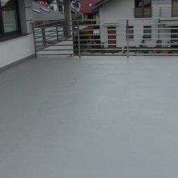 Mikrocement i hydroizolacje - Szary, gładki taras z metalową balustradą, widok na sąsiednie domy z czerwonymi dachami.