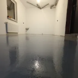 Błyszcząca, szara posadzka żywiczna w pustym pokoju z grzejnikiem pod oknem i rurami pod sufitem. Widok na gładką powierzchnię, odbijającą światło.