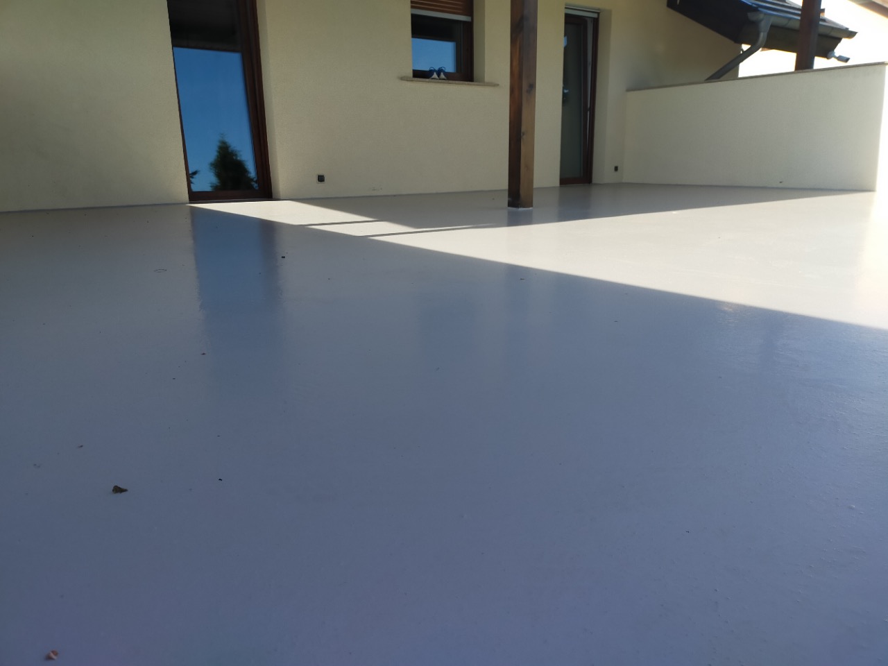 Jasnoszara, błyszcząca powierzchnia tarasu z widocznymi drzwiami balkonowymi, słupem konstrukcyjnym i fragmentem ściany w tle, oświetlona padającym światłem słonecznym.