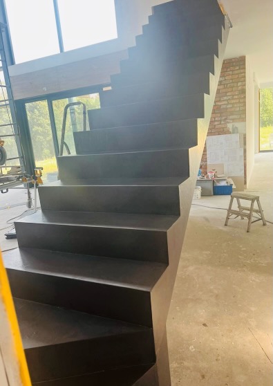 Nowoczesne, ciemnoszare schody wewnętrzne o minimalistycznym designie, z widocznym wykończeniem w stylu industrialnym i surowym betonem na podłodze w tle.