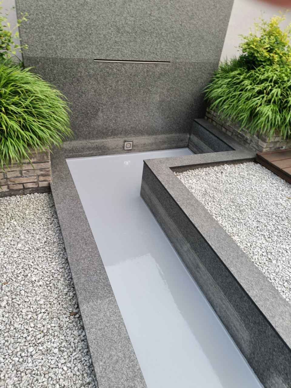 Nowoczesny taras z szarego kamienia i jasną, gładką powierzchnią, otoczony dekoracyjnym żwirem i zielenią, z elementami wodnymi w tle.