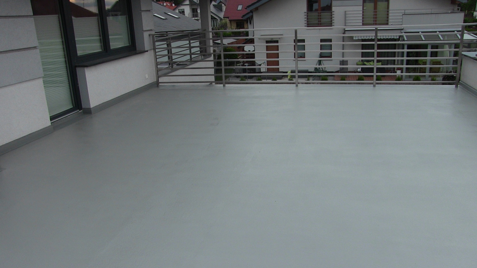 Szary, gładki taras z metalową balustradą, widok na sąsiednie domy z czerwonymi dachami.