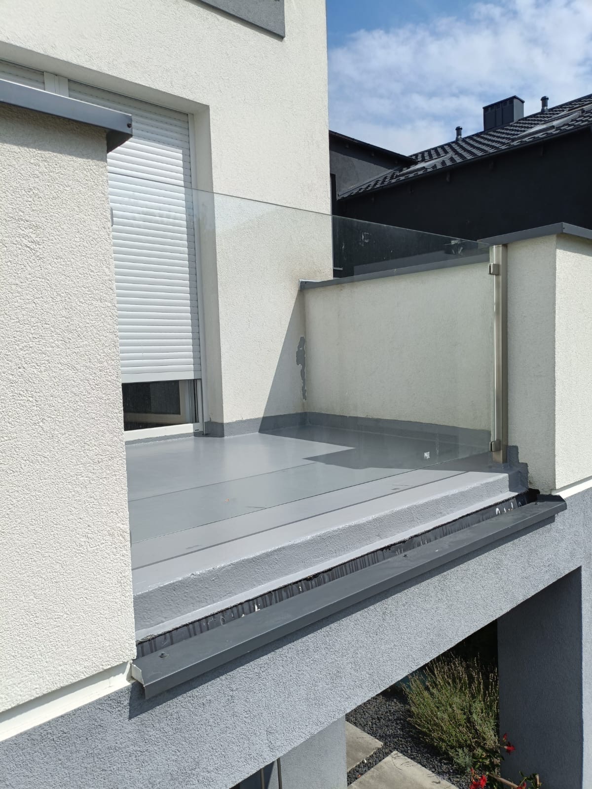 Nowoczesny balkon z szarą, gładką posadzką i szklaną balustradą, widok z zewnątrz na fragment elewacji z żaluzjami.