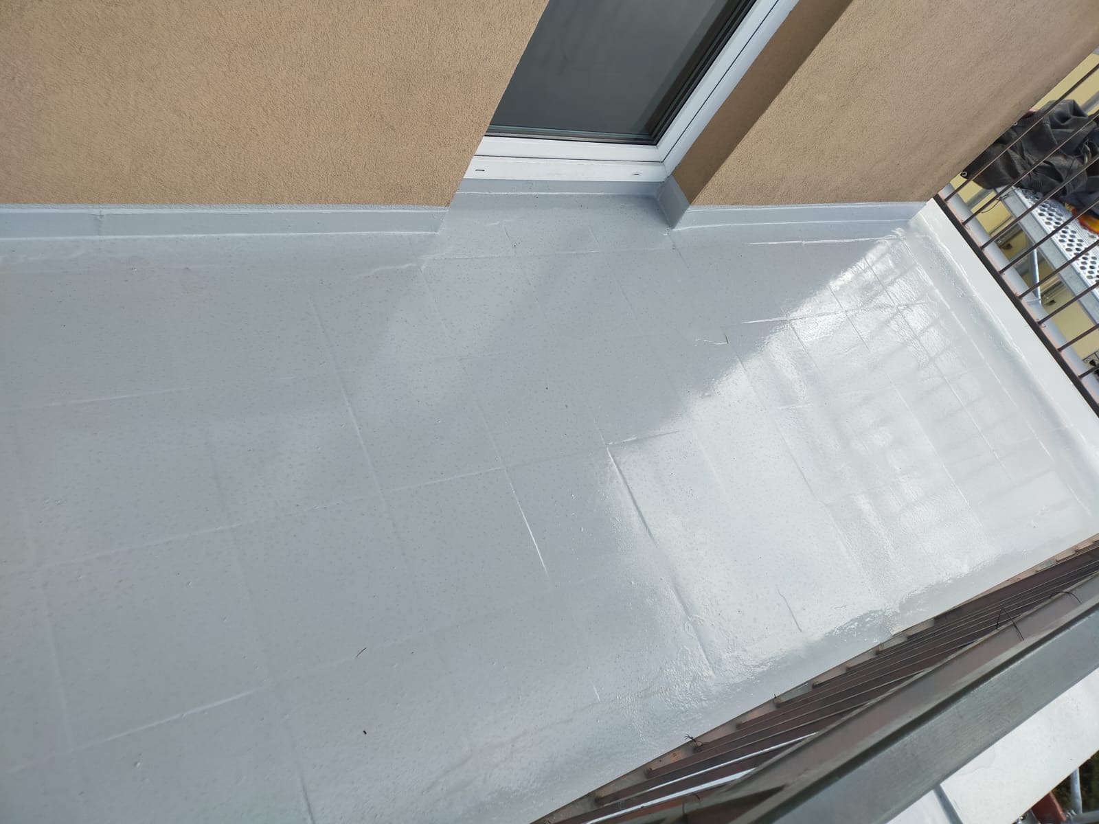 Świeżo wykonana, jasnoszara powierzchnia balkonu z widocznymi liniami podziału na kwadraty, prawdopodobnie płytki, z oknem dachowym w tle i fragmentem metalowej balustrady.