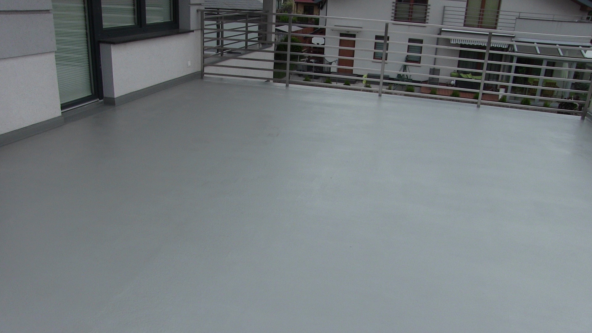 Szary, gładki balkon z metalową balustradą, widok na budynki mieszkalne w tle.