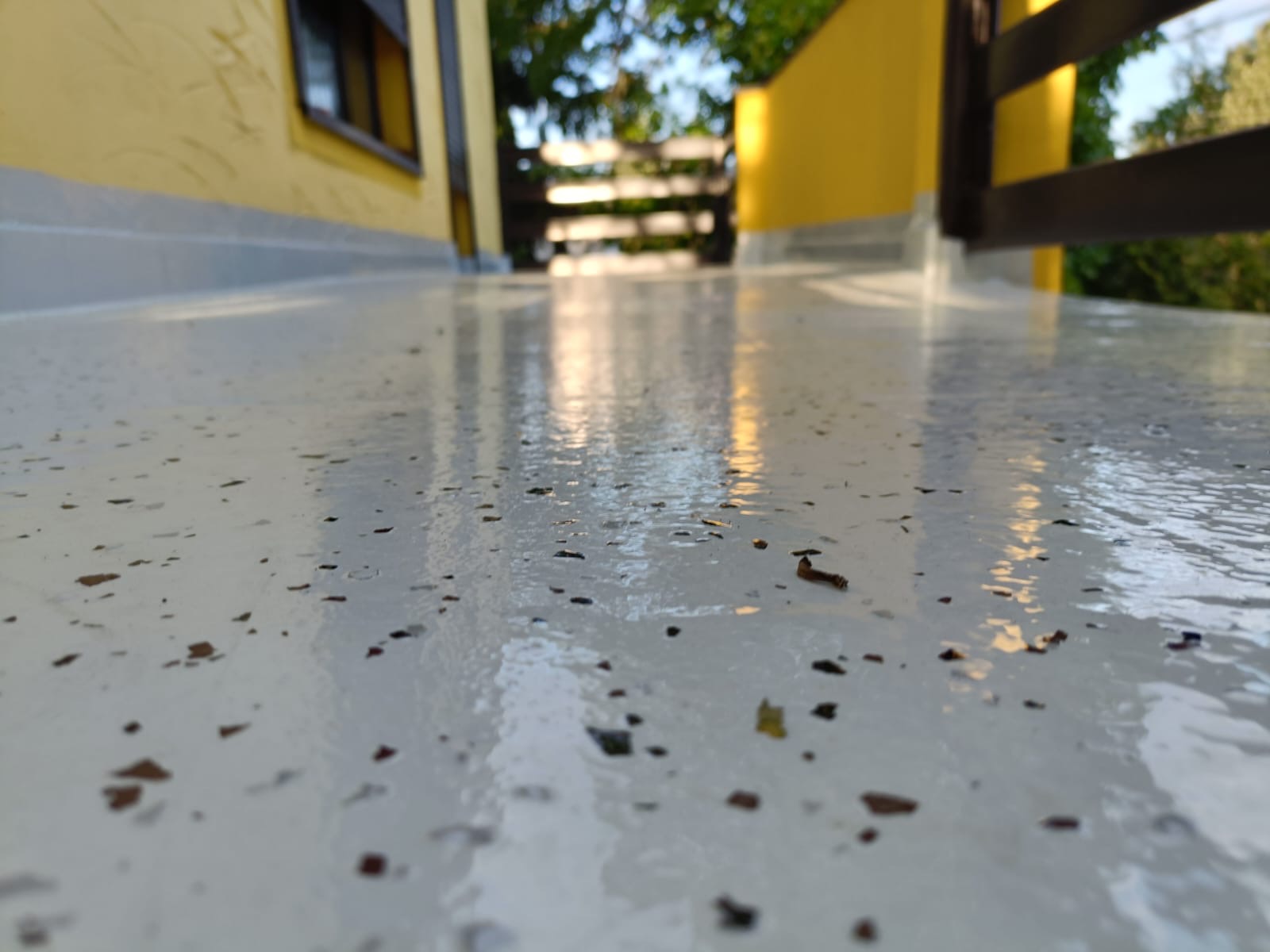 Błyszcząca, jasna posadzka żywiczna na balkonie z widocznymi, drobnymi elementami dekoracyjnymi. W tle żółta elewacja budynku i drewniana balustrada.