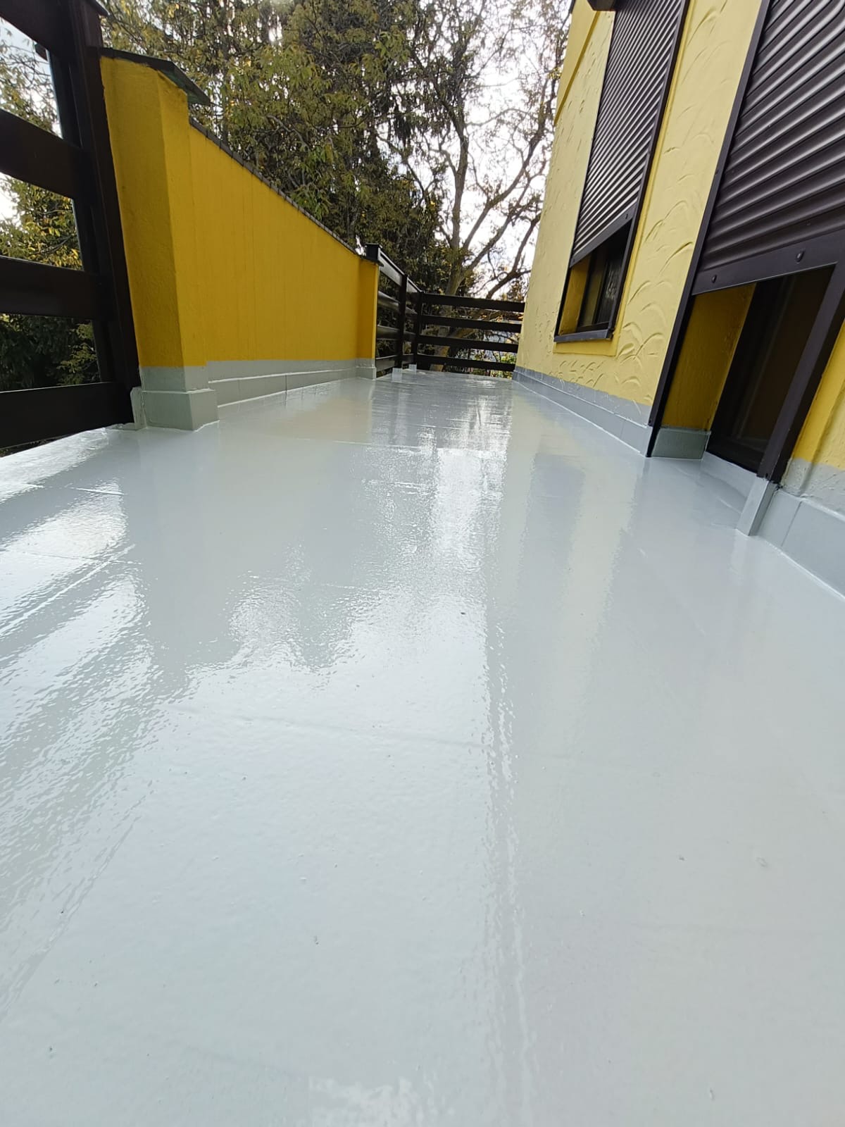 Biała, lśniąca posadzka na balkonie z żółtymi ścianami i drewnianą balustradą. Widoczne refleksy światła na gładkiej powierzchni. Narożne ujęcie.