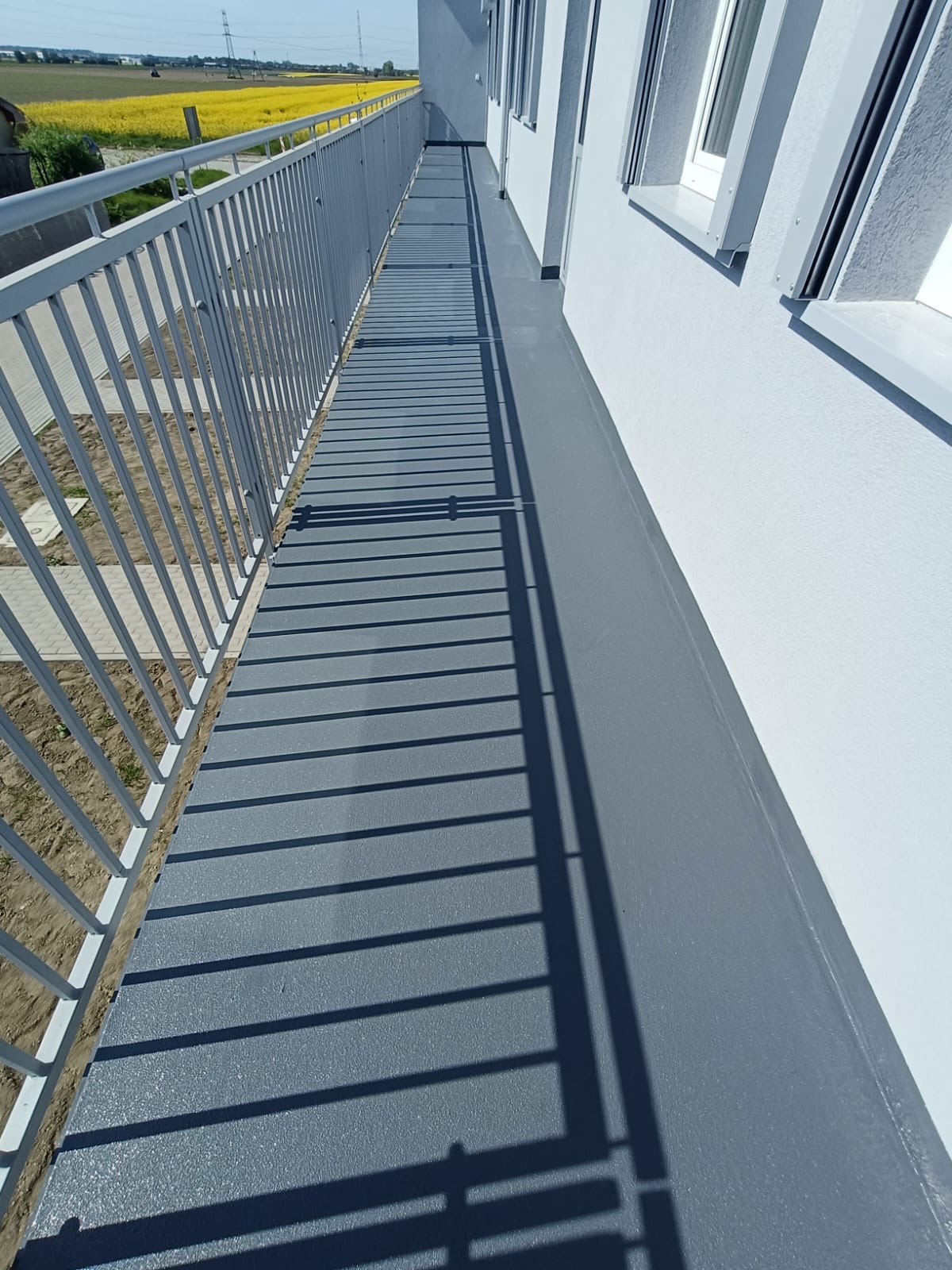 Szary balkon z metalową balustradą, widok z góry. Świeżo położona, gładka posadzka żywiczna odbija promienie słońca. W tle żółte pole i błękitne niebo.