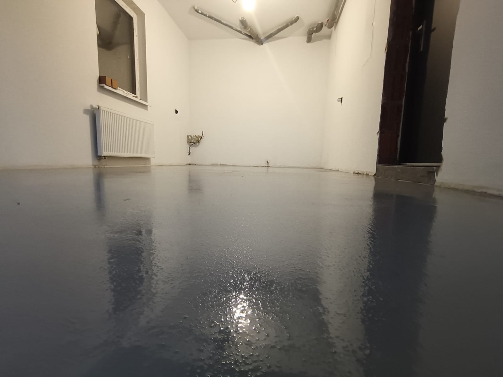 Błyszcząca, szara posadzka żywiczna w pustym pokoju z grzejnikiem pod oknem i rurami pod sufitem. Widok na gładką powierzchnię, odbijającą światło.