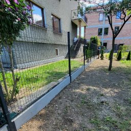 Tomasz Brzeziński - Nowoczesne ogrodzenie panelowe z podmurówką betonową oddzielające trawnik od przygotowanego terenu pod nasadzenia, w tle dom jednorodzinny z balkonem.