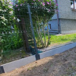 Tomasz Brzeziński - Nowoczesne ogrodzenie panelowe z podmurówką betonową, widoczne zarośla i kwitnący krzew w tle, teren przygotowany do zagospodarowania.