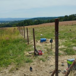 Tomasz Brzeziński - W trakcie budowy ogrodzenia z drewnianych słupków na nierównym terenie, widoczne narzędzia i materiały budowlane, w tle pagórkowaty krajobraz.