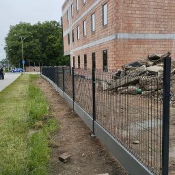 Tomasz Brzeziński - Nowoczesne, czarne ogrodzenie panelowe z betonową podmurówką, ciągnące się wzdłuż placu budowy z widocznymi gruzami i budynkiem w trakcie wznoszenia, z przechodniami na chodniku w oddali.