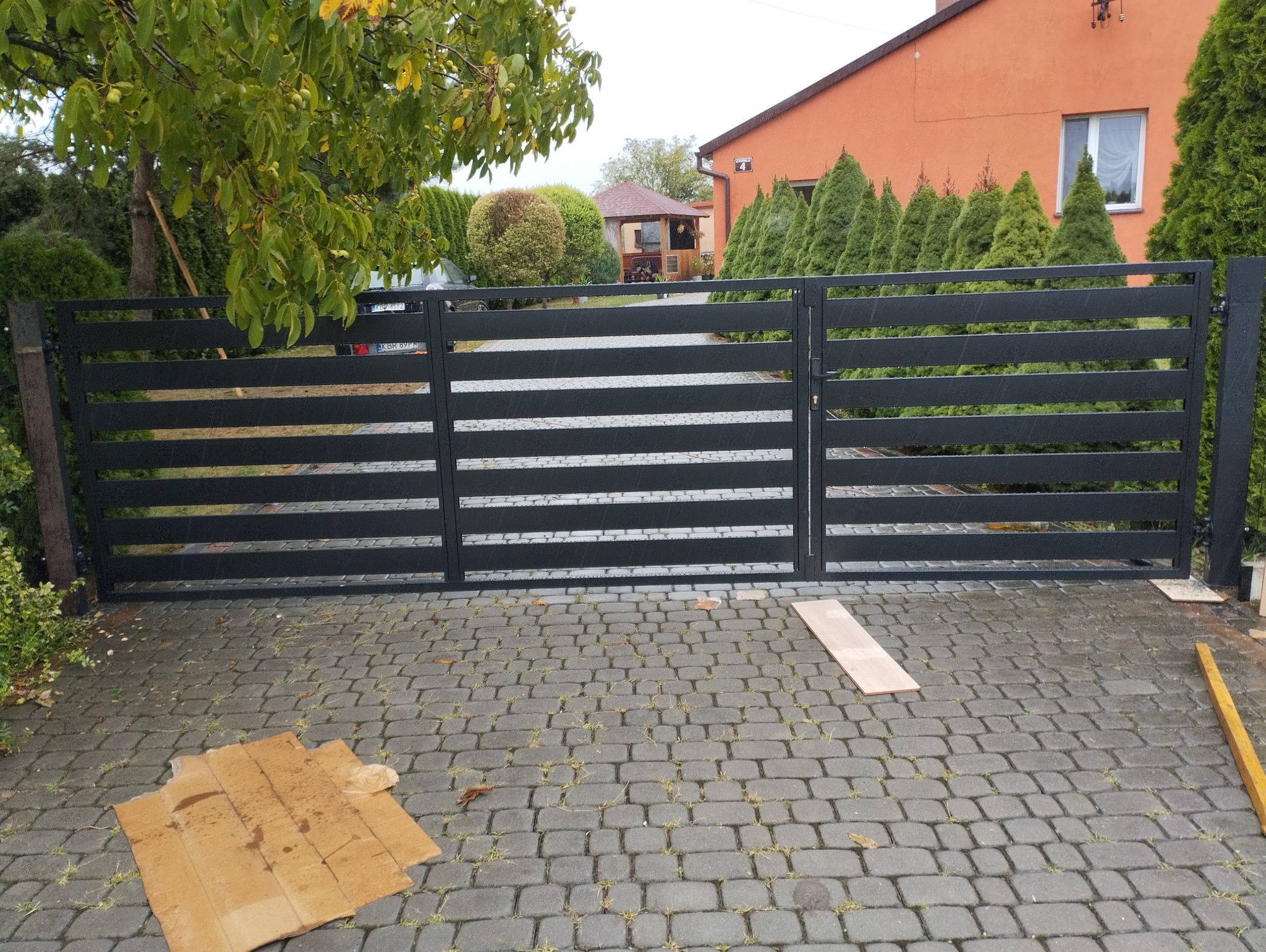 Nowoczesna, pozioma brama metalowa w kolorze grafitowym, z widocznym mechanizmem zamka, zamontowana na posesji z kostki brukowej, z widocznymi elementami montażowymi na podłożu.