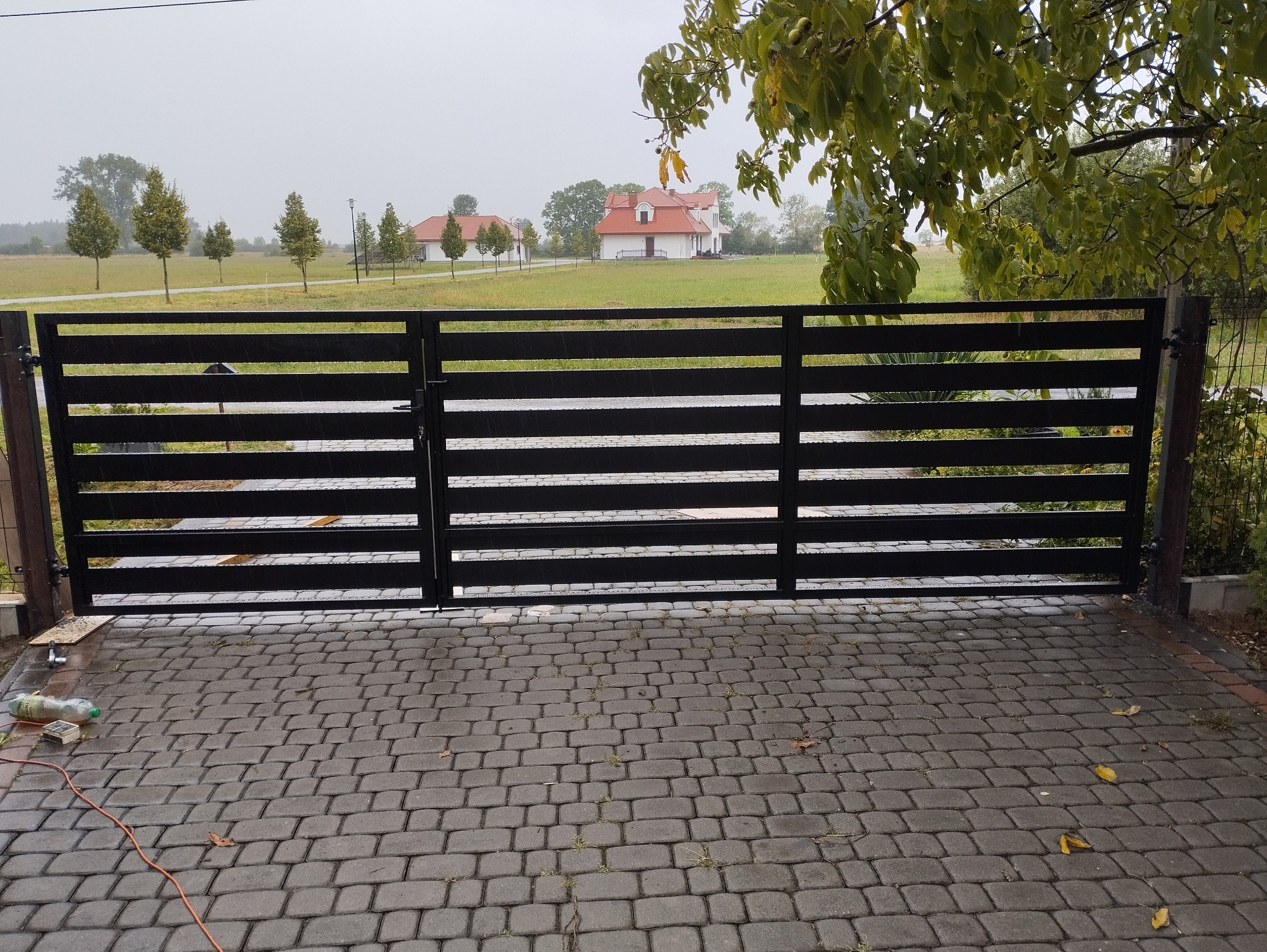 Nowoczesna brama wjazdowa z poziomych, czarnych profili metalowych, na tle domu z czerwoną dachówką i zielonego krajobrazu w pochmurny dzień.
