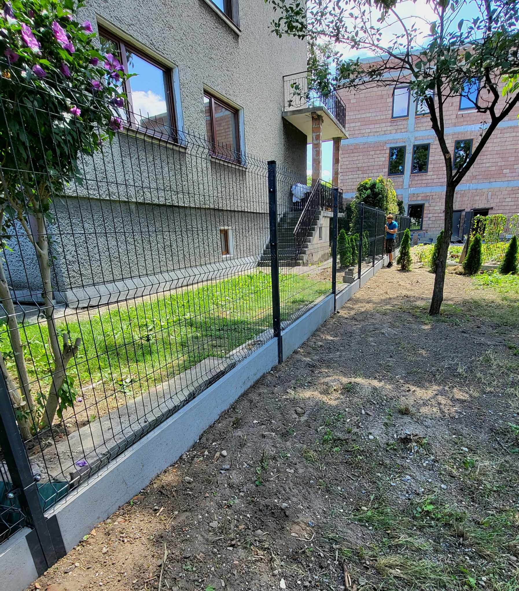 Nowoczesne ogrodzenie panelowe z podmurówką betonową oddzielające trawnik od przygotowanego terenu pod nasadzenia, w tle dom jednorodzinny z balkonem.