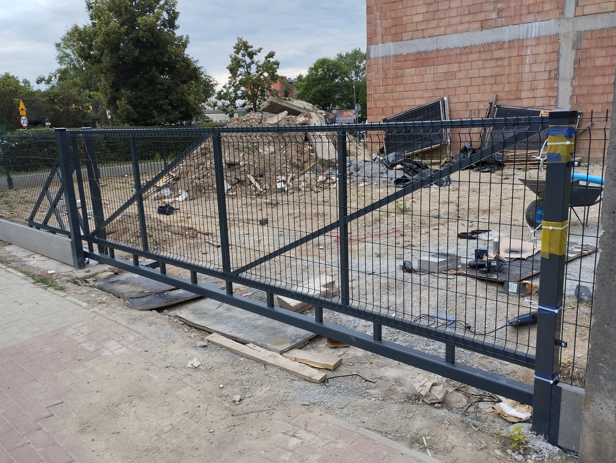 Suwana brama panelowa w trakcie montażu; widoczne narzędzia, gruz i niedokończony budynek w tle.