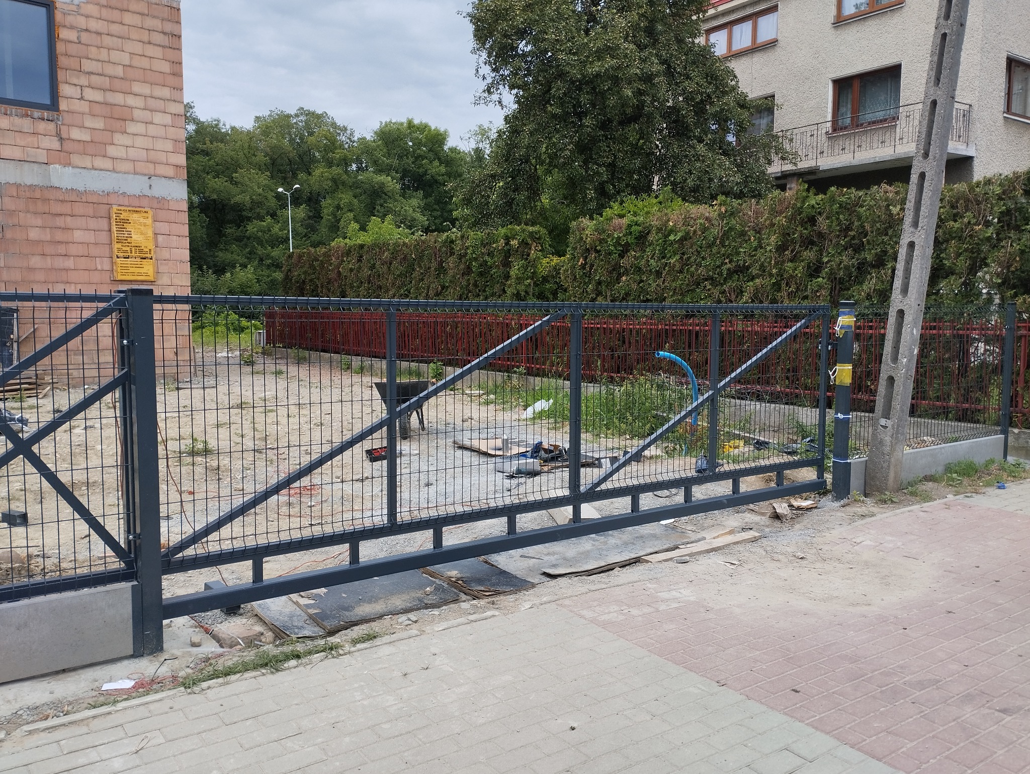 Częściowo zamontowana brama z siatki metalowej w trakcie instalacji, widoczne narzędzia i materiały budowlane na ziemi, w tle żywopłot i budynek.