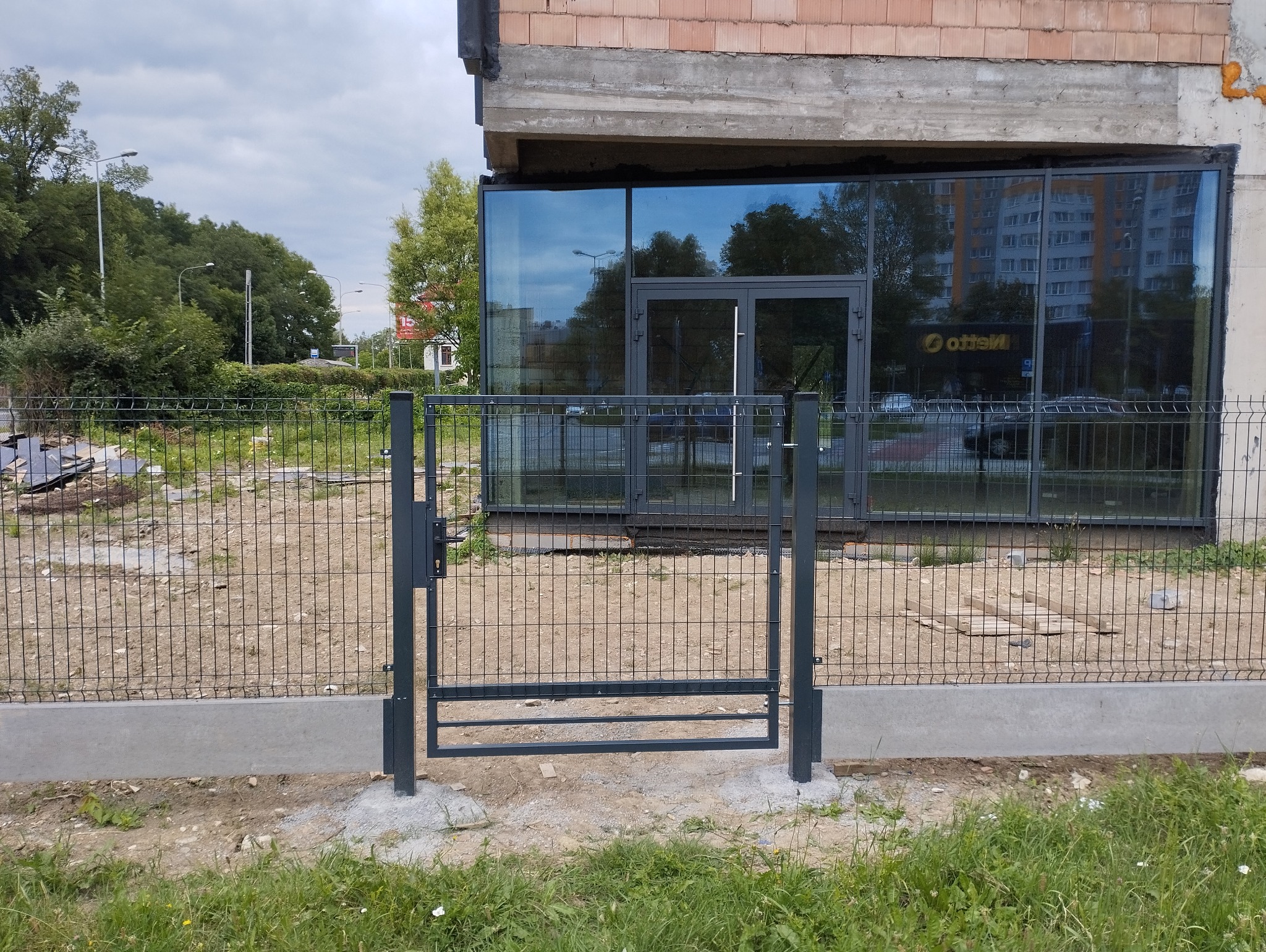 Szara, metalowa furtka panelowa z zamkiem, zamontowana na betonowej podmurówce, widok na budynek z przeszkloną fasadą i odbijające się w szybach drzewa i budynki.