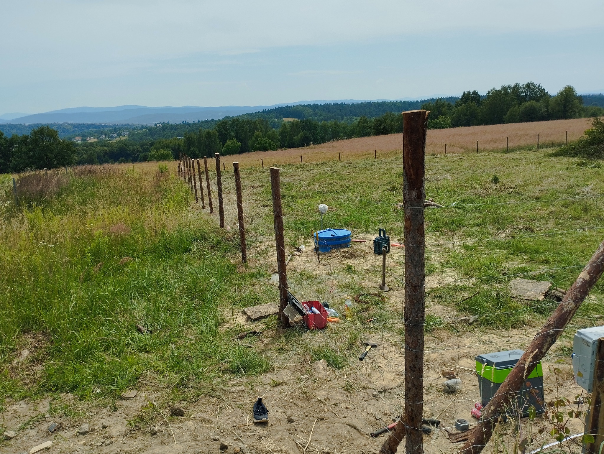 W trakcie budowy ogrodzenia z drewnianych słupków na nierównym terenie, widoczne narzędzia i materiały budowlane, w tle pagórkowaty krajobraz.