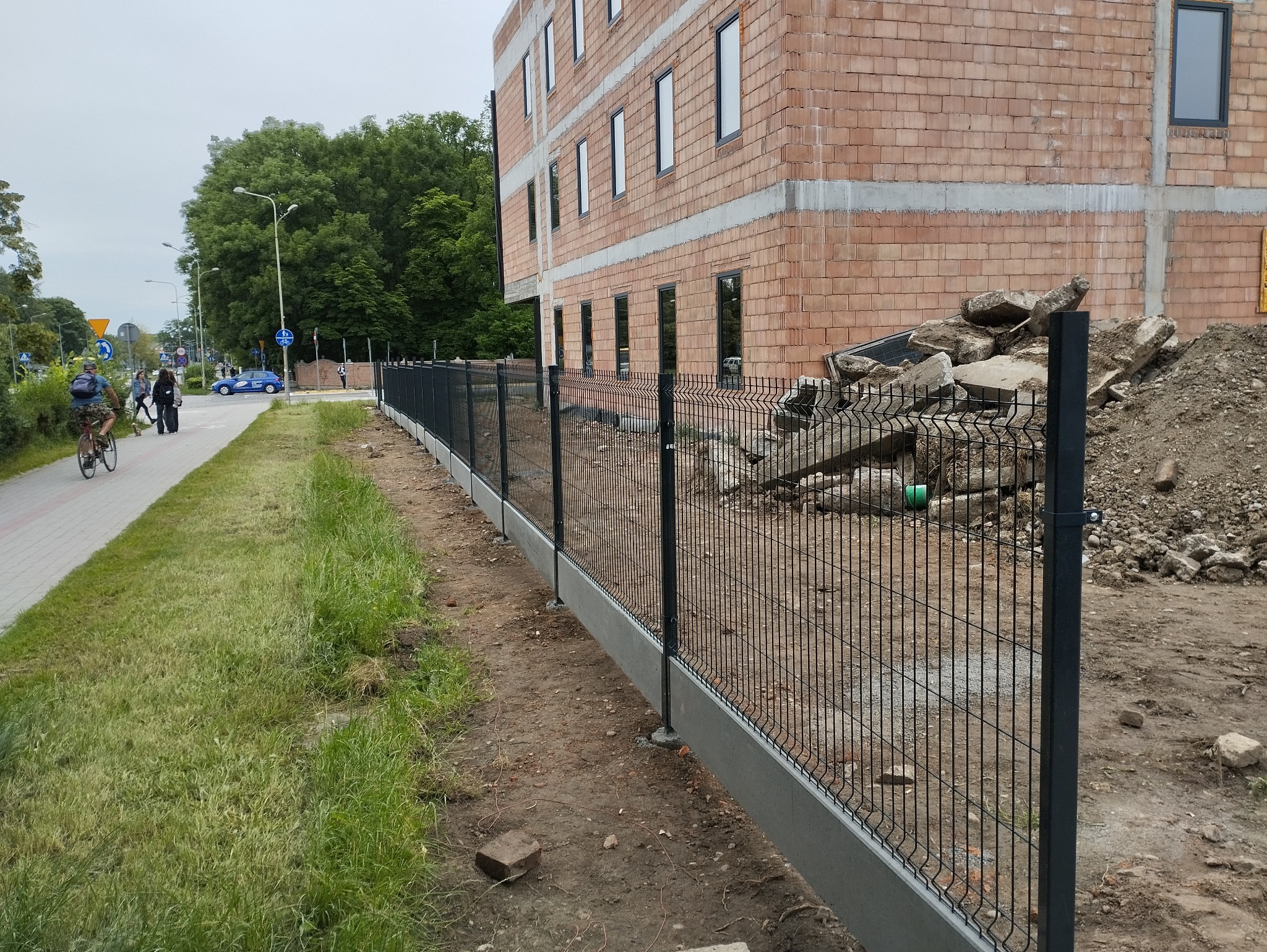 Nowoczesne, czarne ogrodzenie panelowe z betonową podmurówką, ciągnące się wzdłuż placu budowy z widocznymi gruzami i budynkiem w trakcie wznoszenia, z przechodniami na chodniku w oddali.
