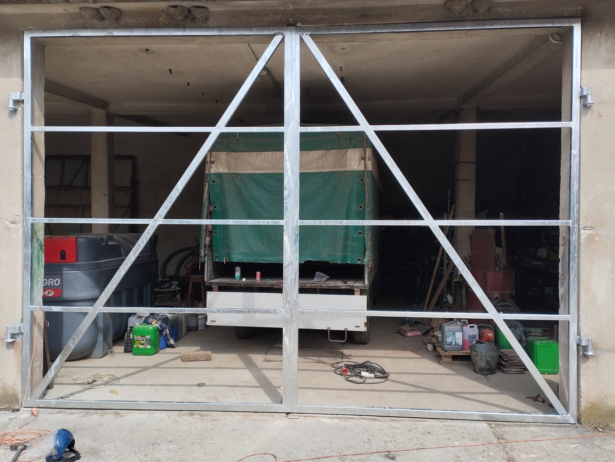 Surowa, metalowa brama garażowa z ukośnymi wzmocnieniami, widok na wnętrze garażu z zaparkowaną ciężarówką z plandeką, w tle liczne przedmioty i pojemniki.
