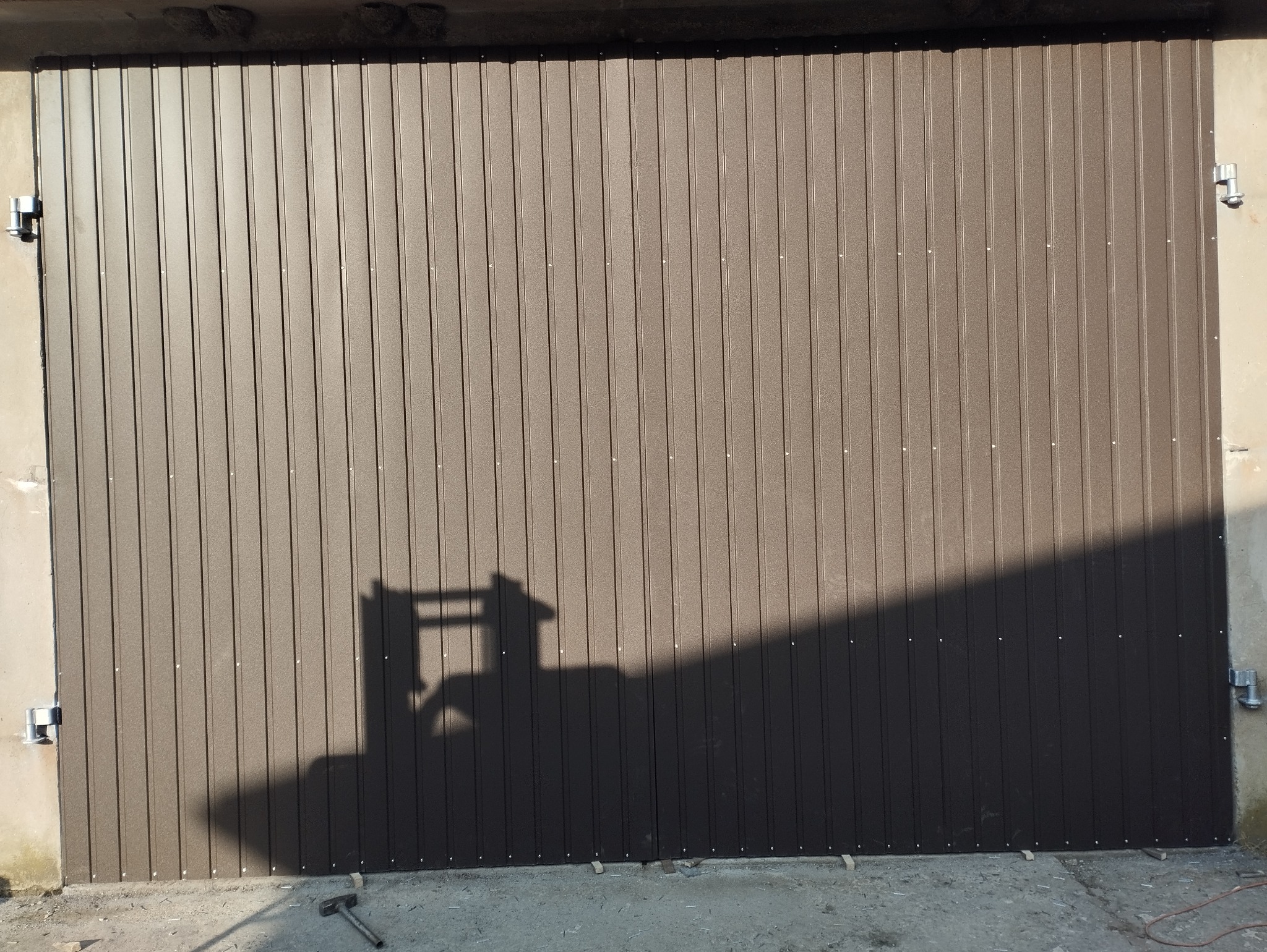 Brązowa brama garażowa segmentowa z widocznym cieniem drabiny i młotkiem leżącym na posadzce.