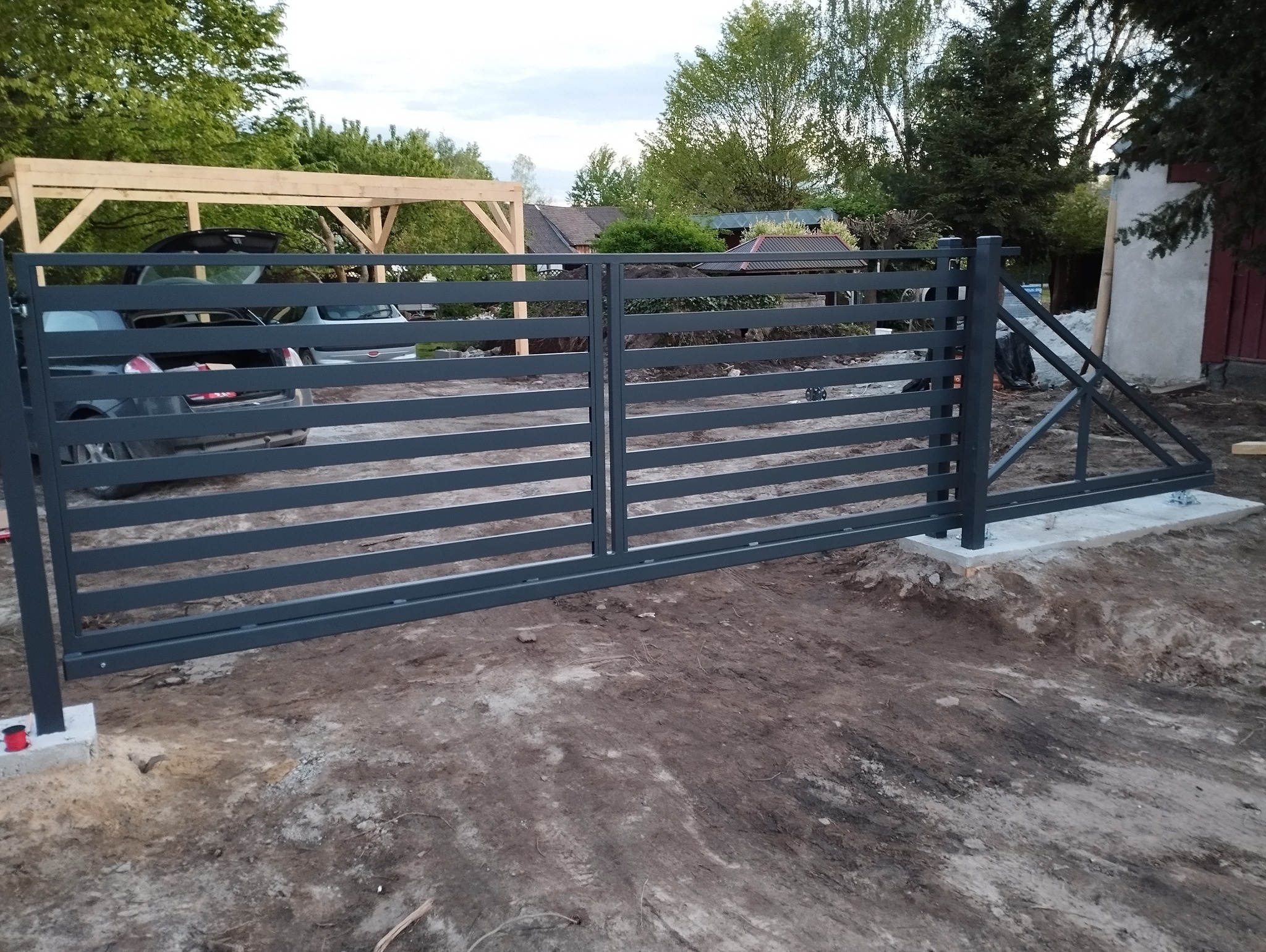 Nowoczesna, pozioma brama wjazdowa z metalowymi przęsłami, szara, z widocznymi betonowymi fundamentami, na tle częściowo zagospodarowanego terenu z drzewami i zabudowaniami.