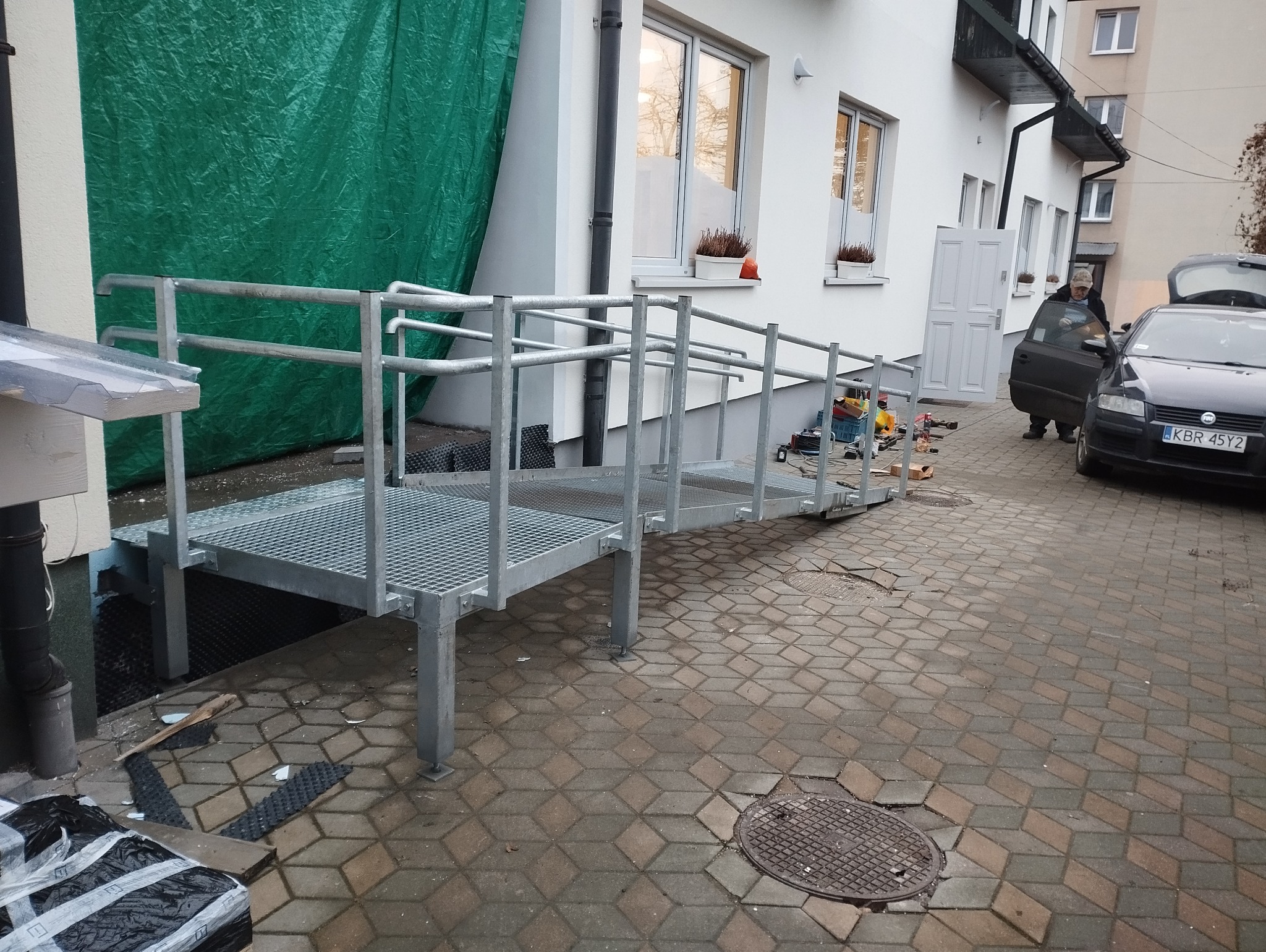 Metalowa rampa dla niepełnosprawnych, prowadząca do budynku, z widocznymi narzędziami i samochodem w tle.