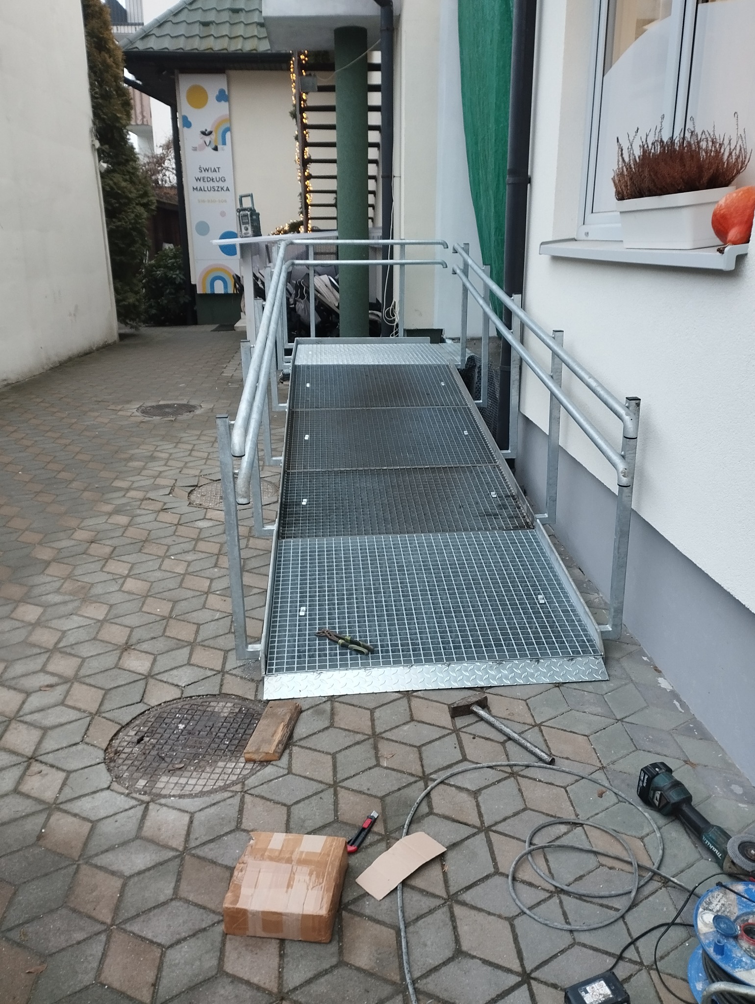Zewnętrzna rampa dla wózków inwalidzkich z metalową kratą i poręczami, obok narzędzia pracy, paczka i właz kanalizacyjny na kostce brukowej.